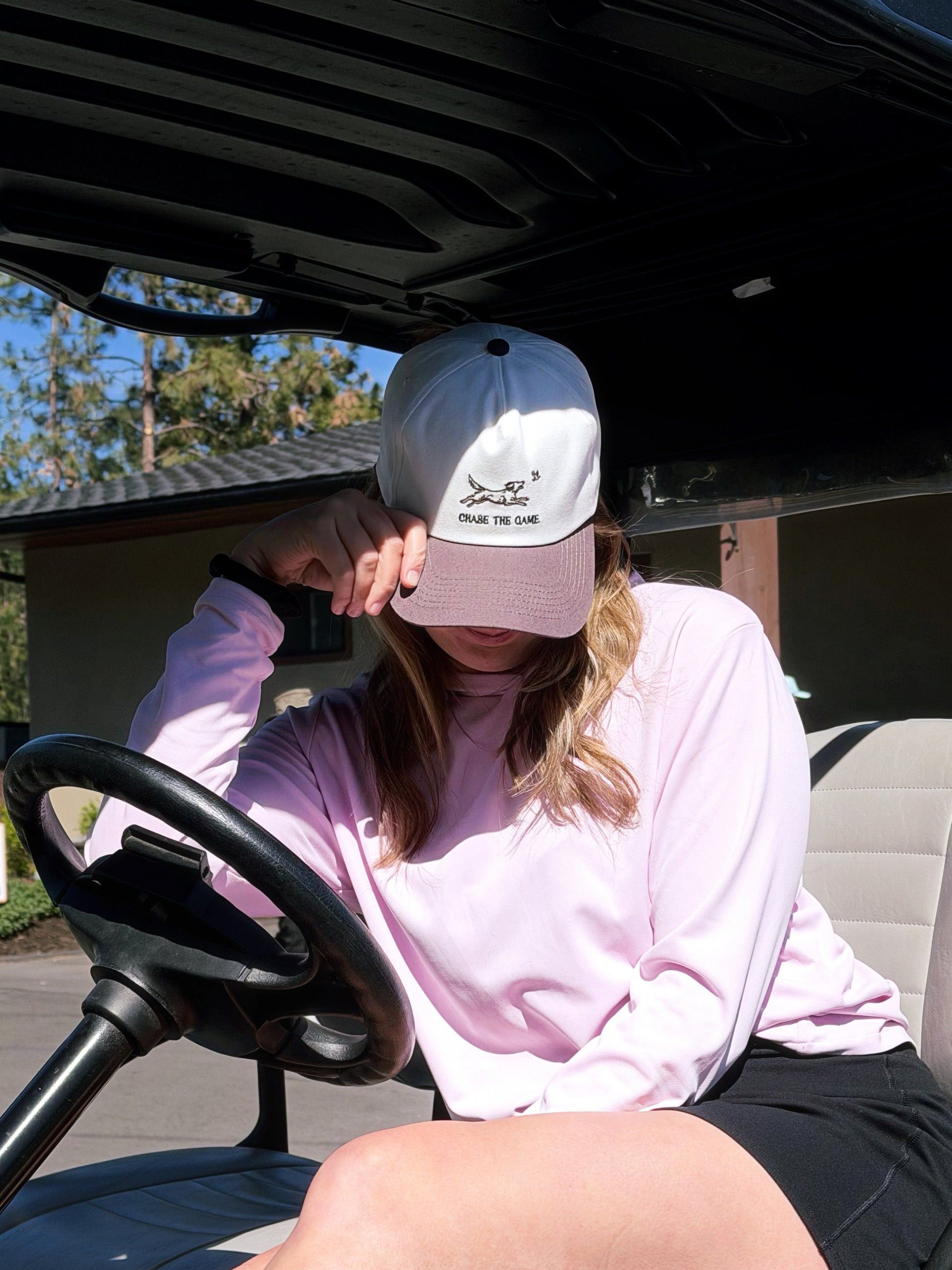 Person sitting in a golf cart wearing a pink cap and light purple sweatshirt.