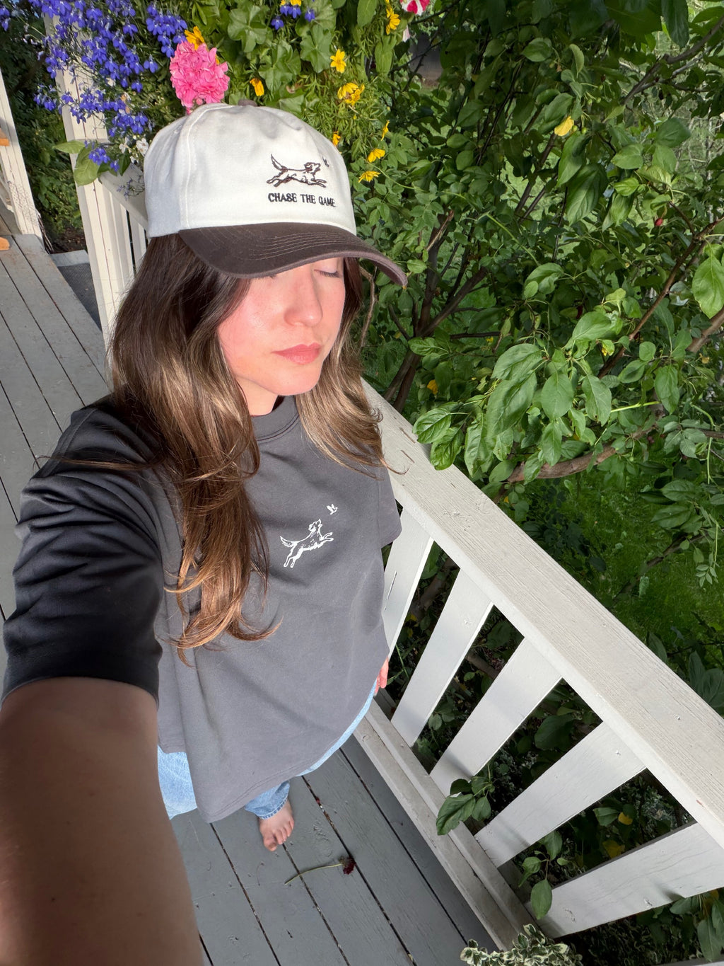 A woman takes a selfie on a wooden deck wearing a white and brown baseball cap with the Quincy Logo and text 'Chase The Game' printed on the front, along with a gray t-shirt